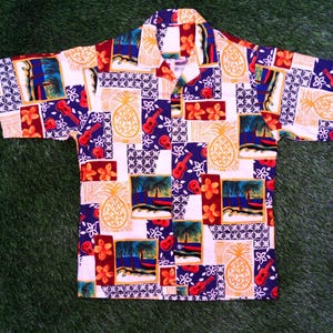May include: Short-sleeved button-down shirt with a colorful tropical print. The design features pineapples, guitars, flowers, and beach scenes in red, blue, yellow, and white. The shirt is laid flat on a green surface.