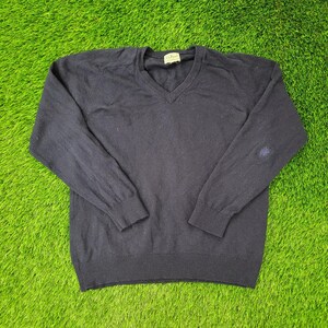 May include: A navy blue V-neck sweater with long sleeves. The sweater has a ribbed hem and cuffs. A small patch is visible on the left elbow. The sweater is lying on a green surface.