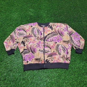 May include: A tan zip-up jacket with a floral pattern in shades of purple, pink, and brown. The jacket has black cuffs, a black hem, and a black collar. The floral design includes stylized flowers and geometric shapes.