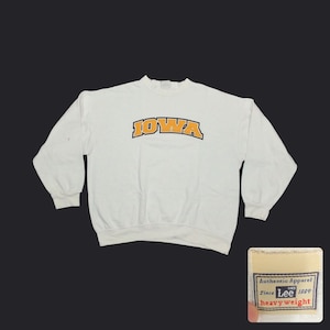 May include: White crew neck sweatshirt with the word "IOWA" in gold and black lettering across the chest. The sweatshirt has long sleeves and a ribbed neckline and cuffs. A tag that says "Authentic Apparel Since Lee 1889 heavy weight" is in the bottom right corner.