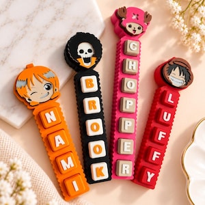 May include: Four vertical silicone cable protectors featuring anime character heads at the top and raised letter blocks spelling NAMI, BROOK, CHOPPER, and LUFFY. The protectors are orange, black, pink, and red, each with a matching character topper.