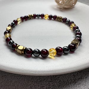 May include: A beaded bracelet featuring a mix of dark red, yellow, and metallic beads. The bracelet includes round and square beads in various sizes. The beads are strung together on a flexible cord, creating a stylish accessory.