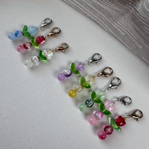 May include: A collection of seven decorative charms, each featuring a metal lobster clasp, a green glass leaf bead, and two additional glass beads in various colors including blue, pink, clear, purple, yellow, and red.