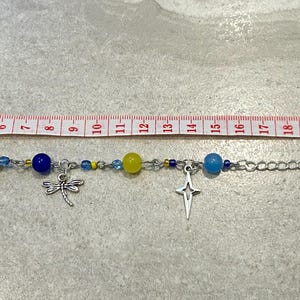 May include: A silver chain bracelet with blue, yellow, and clear beads, and silver charms. The bracelet is approximately 9 inches long. Charms include a spiral, a dragonfly, and a cross.