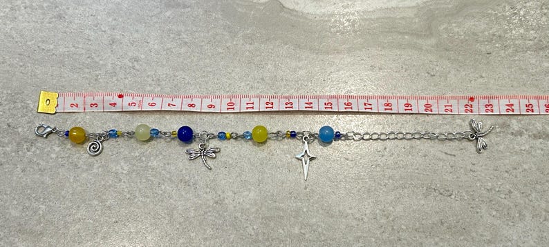 May include: A silver chain bracelet with blue, yellow, and clear beads, and silver charms. The bracelet is approximately 9 inches long. Charms include a spiral, a dragonfly, and a cross.