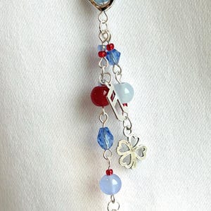 Puede incluir: Un dije de plata con una piedra azul en forma de corazón, cuentas rojas y azules, un trébol de cuatro hojas y una corbata de la suerte. El dije cuelga de una cadena de plata y presenta una mezcla de cuentas redondas y facetadas.