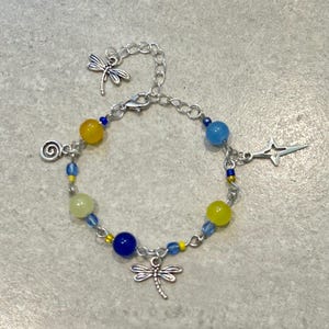 May include: A silver-toned bracelet with a dragonfly charm and various beads in yellow, blue, and green. The bracelet features a chain design with a lobster clasp and additional charms, including a star and a spiral.
