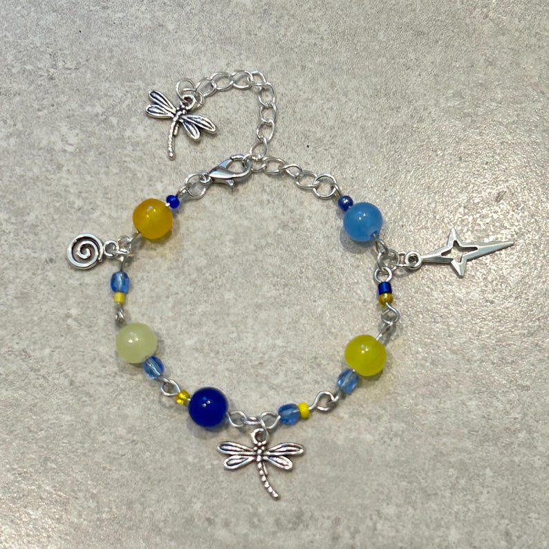 May include: A silver-toned bracelet with a dragonfly charm and various beads in yellow, blue, and green. The bracelet features a chain design with a lobster clasp and additional charms, including a star and a spiral.