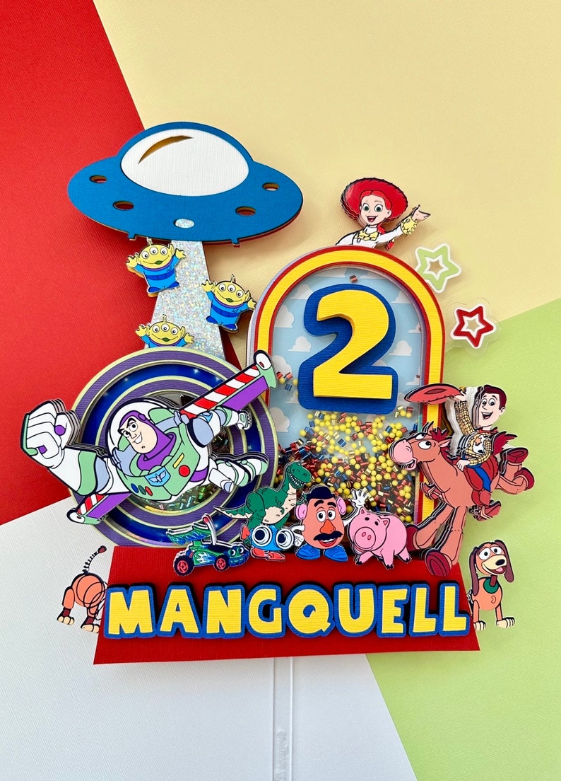 Puede incluir: Adorno de pastel colorido con personajes de Toy Story, incluyendo a Buzz Lightyear y extraterrestres. El dise&ntilde;o incorpora el n&uacute;mero "2" y el nombre "MANGQUELL". El fondo es rojo, amarillo y verde, creando una est&eacute;tica juguetona.