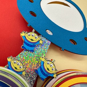 Puede incluir: Una colorida manualidad de papel en capas con tres pines de personajes alien&iacute;genas de dibujos animados. Los alien&iacute;genas tienen tres ojos, trajes azules y piel amarilla. Una nave espacial de papel azul se cierne sobre los alien&iacute;genas.