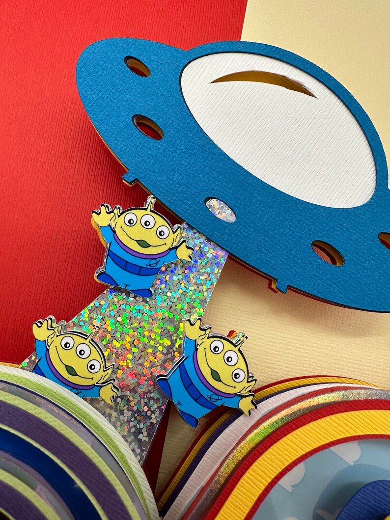 Puede incluir: Una colorida manualidad de papel en capas con tres pines de personajes alien&iacute;genas de dibujos animados. Los alien&iacute;genas tienen tres ojos, trajes azules y piel amarilla. Una nave espacial de papel azul se cierne sobre los alien&iacute;genas.