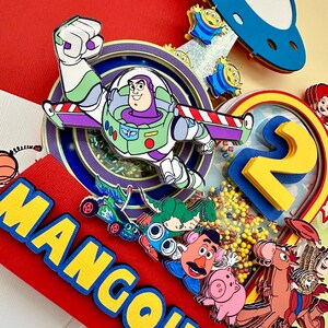 Puede incluir: Una colorida manualidad de papel con Buzz Lightyear, extraterrestres y otros personajes de Toy Story. El dise&ntilde;o incluye el nombre "MANGOUELL" en letras amarillas sobre un fondo rojo y el n&uacute;mero "2" con confeti amarillo.