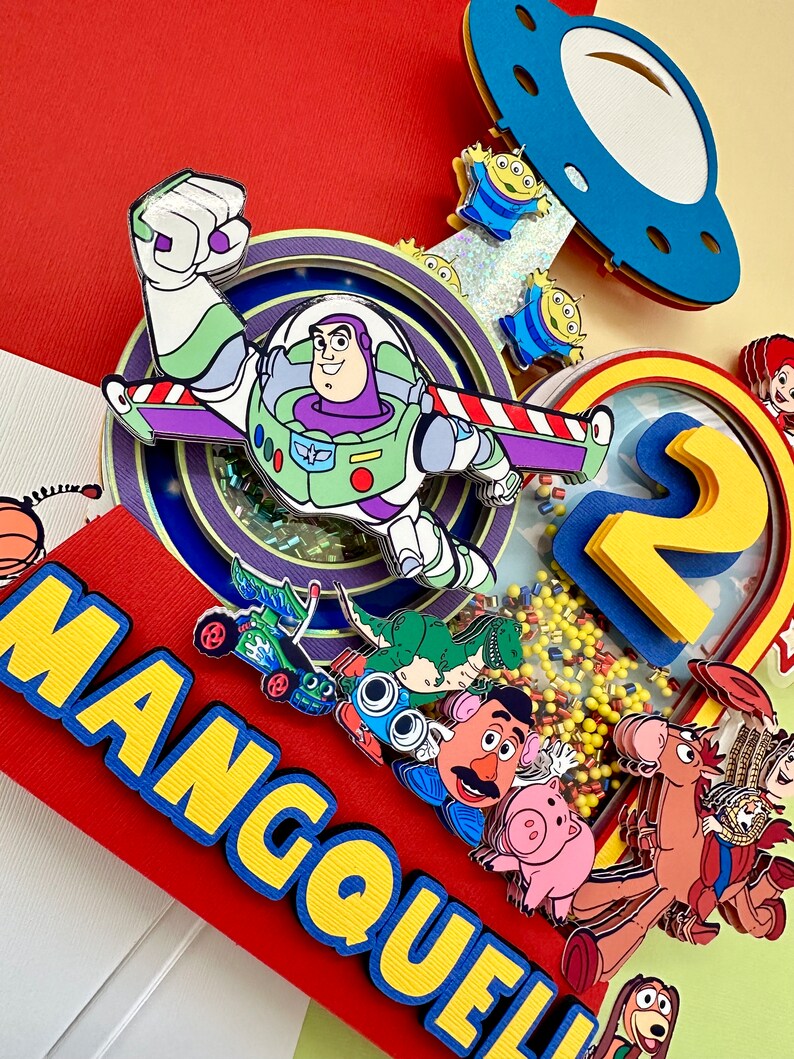 Puede incluir: Una colorida manualidad de papel con Buzz Lightyear, extraterrestres y otros personajes de Toy Story. El dise&ntilde;o incluye el nombre "MANGOUELL" en letras amarillas sobre un fondo rojo y el n&uacute;mero "2" con confeti amarillo.