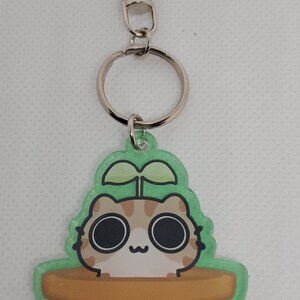 May include: A green and brown keychain featuring a cartoon cat with large eyes peeking out of a flower pot with a paw print. The keychain is attached to a silver key ring.