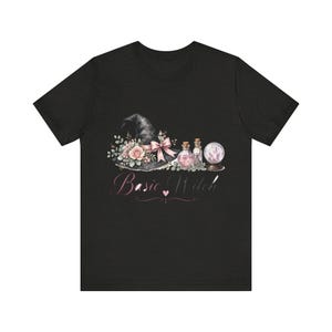 May include: A black t-shirt featuring a graphic of a witch hat adorned with pink roses, a pink ribbon, two small glass potion bottles, and a crystal ball. Below the graphic, the text "Basic Witch" is printed in a pink cursive font with a small heart.