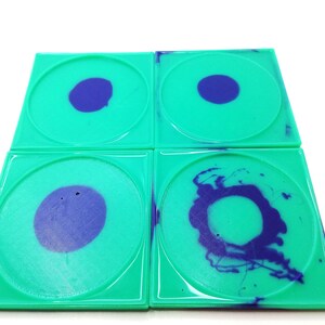 May include: Four square, teal coasters with a circular design. Each coaster has a teal square border and a circular centre with a dark purple design. The designs vary from a solid circle to a more abstract pattern.