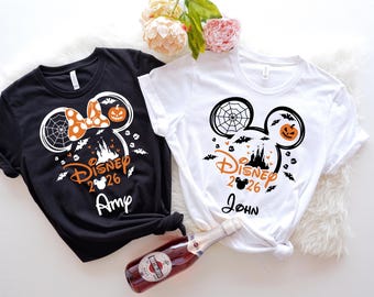 May include: Black and white t-shirt with a Halloween-themed Mickey Mouse design. The design includes a spiderweb, bats, a pumpkin, and the text "Disney 2026". A white t-shirt with a similar design is also shown.