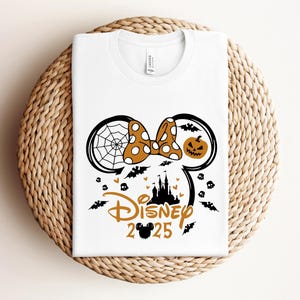 May include: White t-shirt with a Halloween-themed design. The design features a Minnie Mouse silhouette with a spiderweb, pumpkin, bats, and the text "Disney 2025" in orange and black. The shirt is displayed on a woven surface.