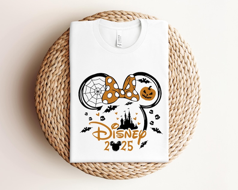 May include: White t-shirt with a Halloween-themed design. The design features a Minnie Mouse silhouette with a spiderweb, pumpkin, bats, and the text "Disney 2025" in orange and black. The shirt is displayed on a woven surface.