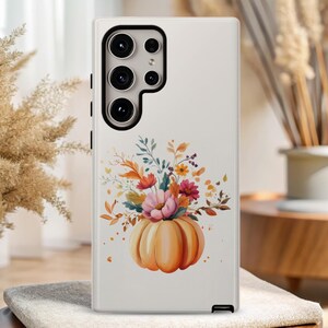 May include: A white phone case with a floral design and a pumpkin. The pumpkin is orange and has a green stem. The flowers are pink, yellow, and orange. The phone case has a camera lens cutout.