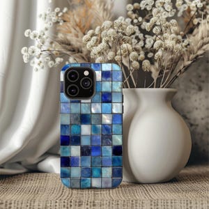 May include: A smartphone case with a mosaic tile design in shades of blue. The case features a square pattern with varying tones of blue, creating a textured appearance. The phone case is next to a white vase with dried flowers.