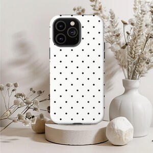 May include: A white phone case with a black polka dot pattern. The case has a camera cutout and is displayed on a concrete pedestal. Decorative white flowers and a vase are in the background.