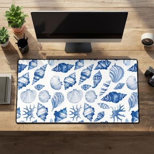 May include: A rectangular desk mat featuring a white background with a repeating pattern of blue seashells. The design includes various shell shapes and sizes, creating a coastal or nautical theme. The mat is placed on a wooden desk, with a computer monitor and other office supplies visible.