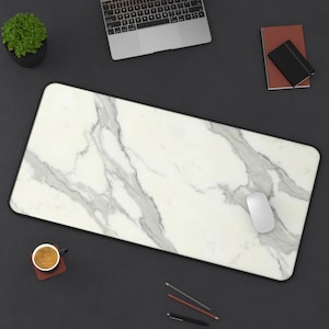 May include: A rectangular white and gray marble-patterned desk mat with a black border. A white computer mouse rests on the mat. A laptop, a small potted plant, a notebook, a cup of coffee, and writing utensils are also visible.