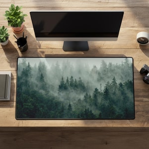 May include: A rectangular desk mat featuring a misty forest scene with tall green trees. The mat is on a wooden desk, with a computer monitor, plants, and other office supplies visible.