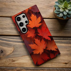 May include: A smartphone case featuring a vibrant autumn leaf design. The case showcases a pattern of red and orange maple leaves, creating a seasonal aesthetic. The phone case is on a wooden surface.