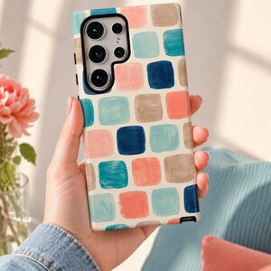 May include: A rectangular smartphone case featuring a repeating pattern of rounded squares in shades of teal, coral, navy blue, and beige against a white background. The case has a black camera cutout area and a small black stylus tip visible at the bottom.
