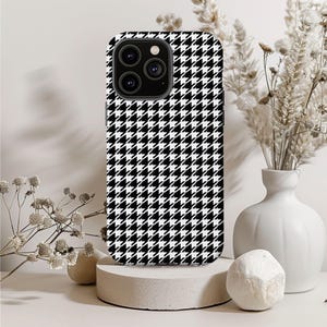 May include: A smartphone case with a black and white houndstooth pattern. The case has a camera cutout and is displayed on a small, round concrete pedestal. The background includes a white vase with dried flowers and decorative elements.
