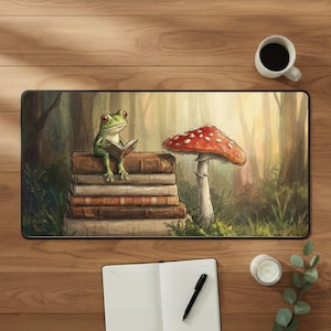 May include: A whimsical mousepad featuring a green frog reading a book atop a stack of books, with a red-capped mushroom nearby. The background depicts a forest scene. A cup of coffee, a notebook, a pen, a candle, and greenery are on the wooden surface.