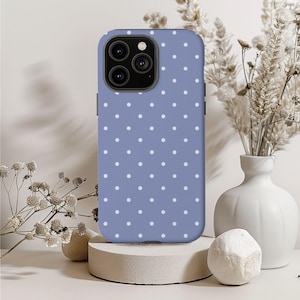 May include: A periwinkle blue phone case with a white polka dot pattern. The case has a camera cutout and is displayed on a concrete pedestal. The background includes a white vase with dried flowers and decorative stones.