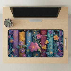 May include: A rectangular mousepad featuring a vibrant illustration of colorful, stacked books adorned with flowers and vines. The books are in shades of purple, yellow, and blue, with a dark background. A succulent and a pen are also visible.