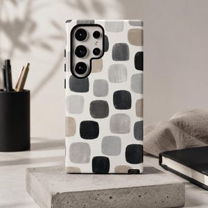 May include: A rectangular smartphone case featuring a repeating pattern of rounded squares in shades of black, gray, and beige against a white background. The case has a large cutout for a multi-lens camera system and a small opening at the bottom corner.