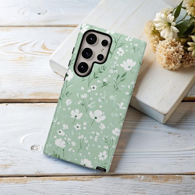 May include: A green phone case with a white floral pattern. The case has a camera cutout on the back.