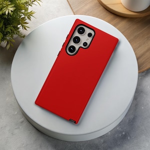 May include: A bright red smartphone case with a multi-lens camera array. The case is rectangular and covers the back and sides of the phone. The phone case is on a white circular surface.