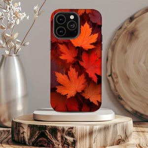 May include: A smartphone case featuring a vibrant autumn leaf design. The case showcases a pattern of red and orange maple leaves, creating a seasonal aesthetic. The case is displayed on a wooden stand.