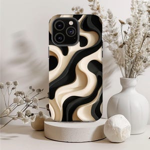 May include: A phone case with a layered design in black, cream, and dark gray. The case has a three-camera system and is displayed on a small, round concrete pedestal. The background includes a white vase with dried flowers and decorative stones.