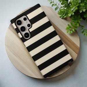 May include: A black and cream striped phone case. The case has a rectangular shape with a camera cutout and is resting on a wooden surface. The case features a bold, horizontal stripe pattern.