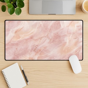 May include: A rectangular mouse pad with a pink marble design. The pad has a black border and a white computer mouse is next to it. A laptop, a notepad, a pen, and a cup of coffee are also on the wooden surface.