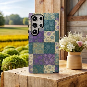 May include: A patterned phone case featuring a patchwork design with floral and paisley motifs in shades of purple, teal, and cream. The case is displayed on a wooden surface with a blurred background of a field and a vase of flowers.