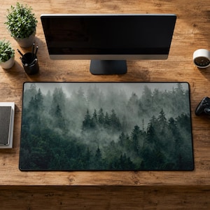 May include: A rectangular desk mat featuring a high-resolution print of a dense, misty forest with dark green pine trees shrouded in thick fog. The mat has a black border and is positioned on a wooden surface in front of a computer monitor.