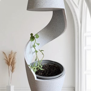 May include: A contemporary, grey, spiral-design planter with a small green plant. The planter has a textured surface and a circular top. The plant pot is dark grey and filled with soil. The background is a white wall and a window.