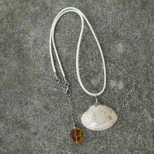 May include: A seashell pendant necklace with a cream-colored cord. The pendant is a natural, off-white seashell with brown markings. A small, amber-colored charm hangs below the shell. The necklace is displayed on a gray surface.
