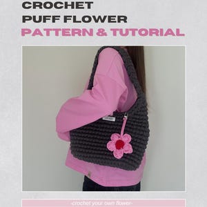 以下が含まれることがあります： ピンクの花のチャームが付いたグレーのクロシェトートバッグ。ピンクのシャツを着た人が肩にかけています。画像の下部に「crochet your own flower」というテキストがあります。
