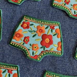Könnte beinhalten: Fünf gestickte Patches in Unterwäscheform mit einem Blumenmuster. Die Patches befinden sich auf einem blauen Jeans-Hintergrund. Die Blumen sind rot, orange und gelb mit grünen Blättern.