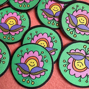 Pode incluir: Uma coleção de patches verdes circulares com um design floral. Cada patch apresenta um sol sorridente no centro, rodeado por pétalas rosa, folhas roxas e pequenos pontos amarelos. Os patches têm uma borda preta.