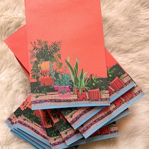 Op de afbeelding: Een stapel koraaloranje notitiekaartjes met een kleurrijke illustratie van potplanten op de voorkant. De illustratie toont een verscheidenheid aan planten in verschillende gekleurde potten met een blauwe en bruine gepatenteerde rand.